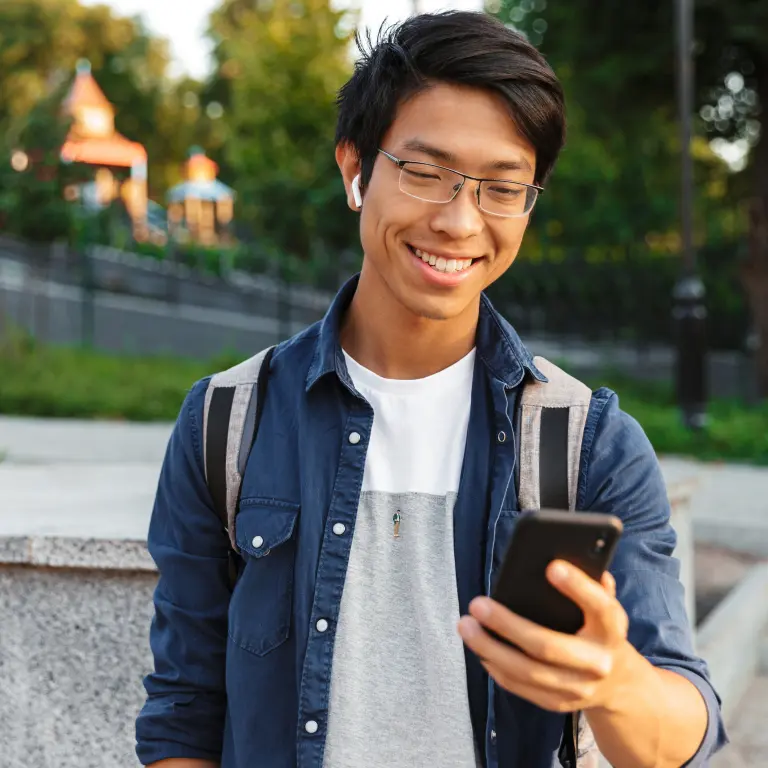 Smiling student wearing glasses and earphones, holding a smartphone outdoors with a backpack on his shoulders.